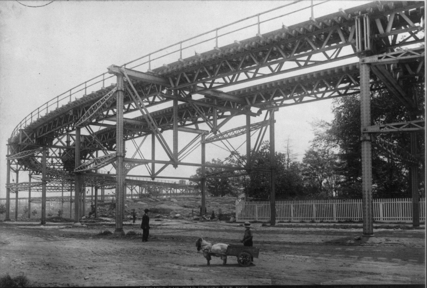 Looking up at the lattice girders and tall columns of the 110th Street curve in 1883, with a man and a child's goat cart on the unpaved street below for scale.