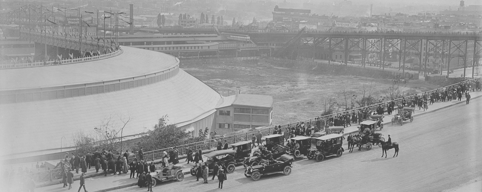 View from the Speedway over the Polo Grounds towards the 155th Street terminal, with the elevated structure's X-braced bents running across the right of the picture and crowds and early motor cars in the foreground.